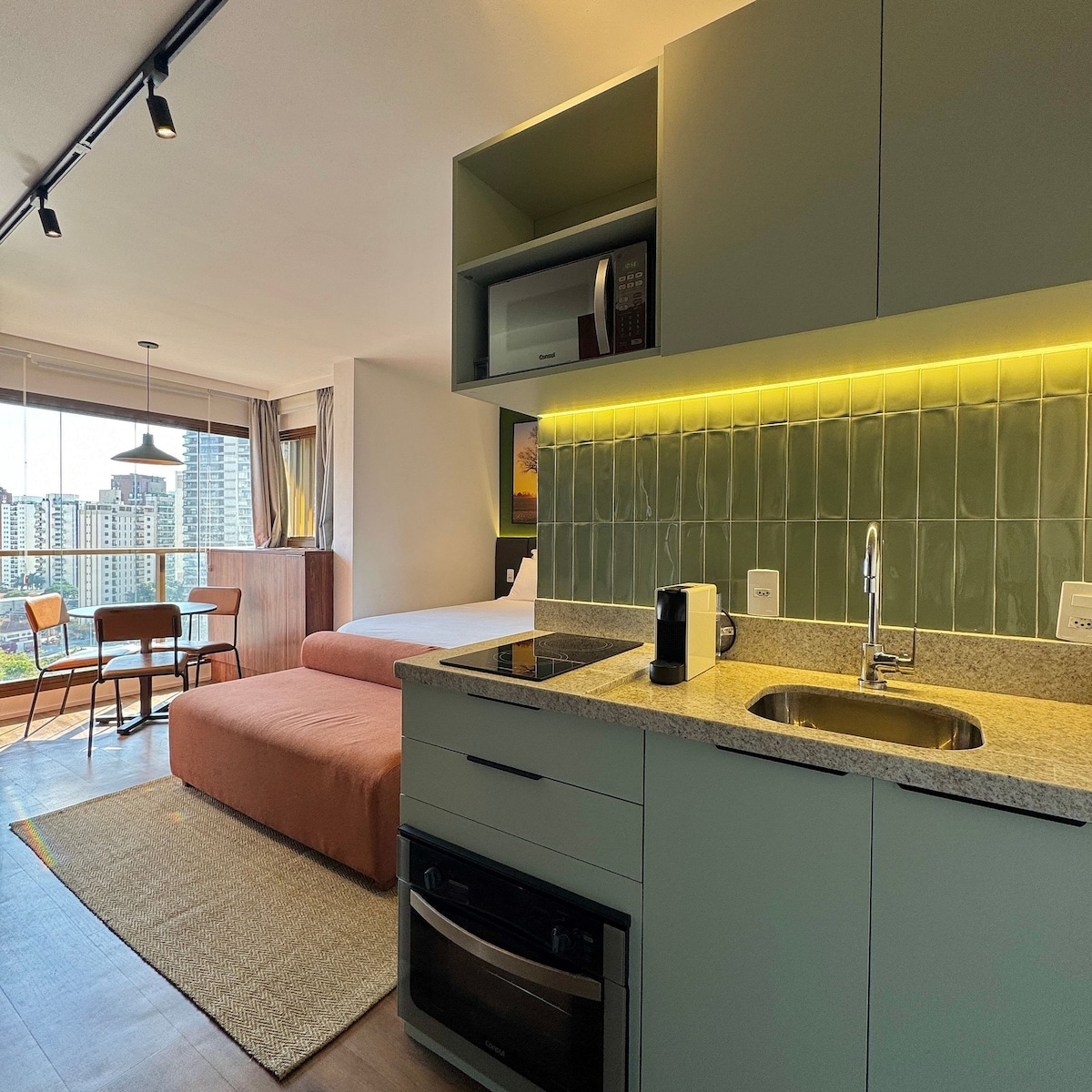 The image presents a well-equipped kitchenette featuring a countertop with a sink and modern appliances, including a microwave and coffee maker. A comfortable seating area with a small dining table and chairs is visible next to a large window, allowing natural light to brighten the space.