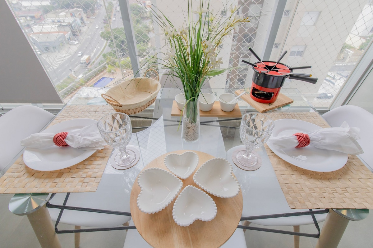 A modern dining table is set with heart-shaped dishes and elegant glassware. A centerpiece features a decorative plant, and a fondue pot adds a unique touch. The background showcases a city view, visible through large windows, enhancing the dining experience.