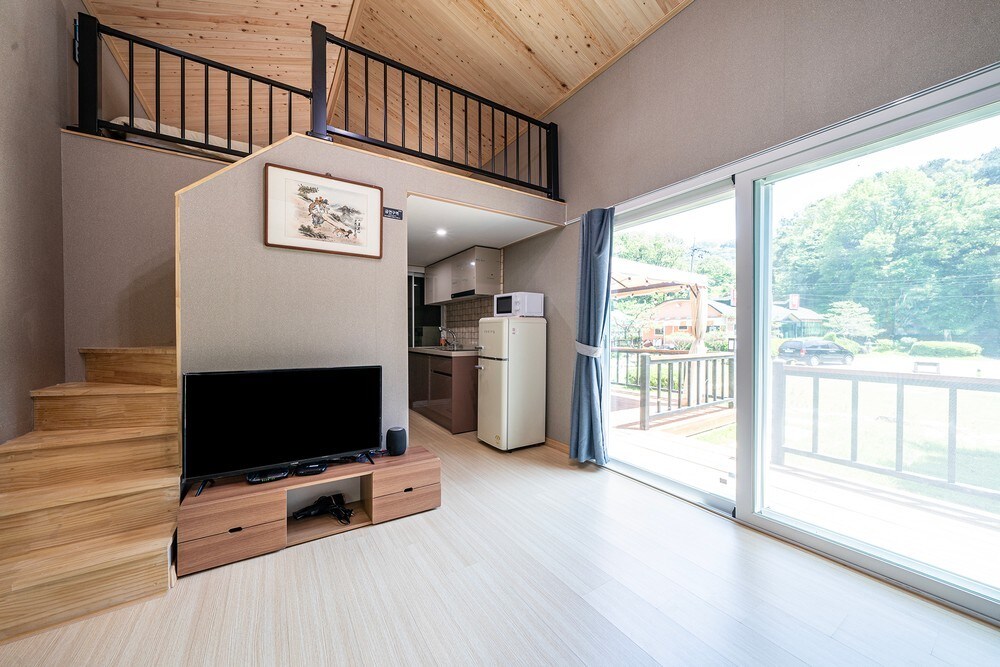 A spacious living area features wooden flooring, a flat-screen TV on a low cabinet, and a clear view through large glass doors leading to an outdoor space. A staircase leads to an upper level, and a kitchenette with modern appliances is visible in the background.