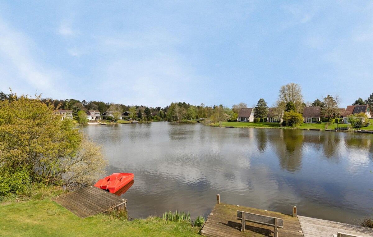 A serene lakeside view showcases calm water reflecting a clear sky, surrounded by lush greenery and residential properties. A wooden dock leads to the water, featuring a bright red boat, inviting relaxation in this peaceful setting.