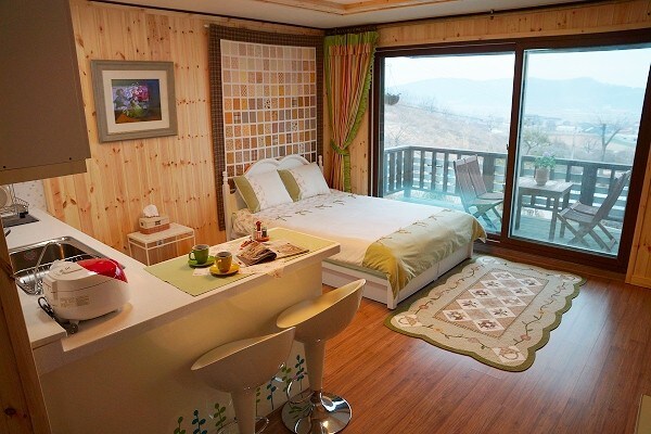 A bright room features a spacious bed with light green bedding, complementing the wooden decor. A small kitchenette with two stools is visible. A large window offers a view of the outdoors, with a balcony set with chairs and a table.