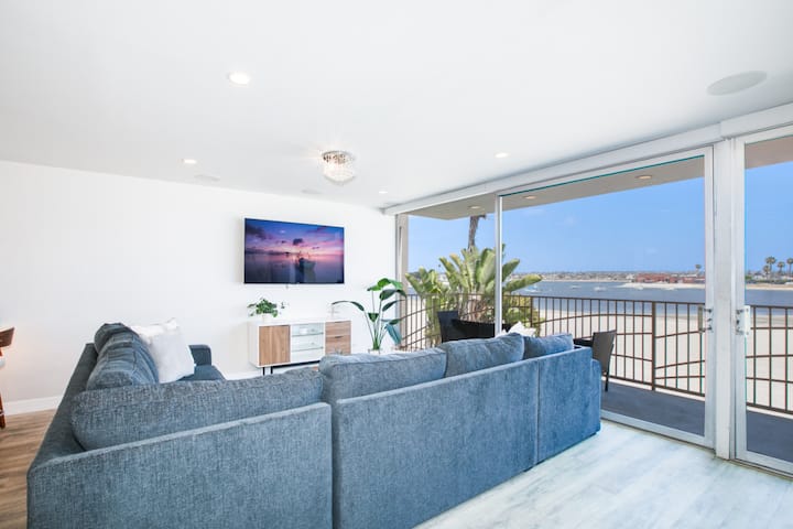 Svetlý a pohodlný obývací priestor s veľkou modrou sekčnou pohovkou orientovanou na televízor s plochou obrazovkou namontovaný na elegantnej mediálnej konzole. Sklenené dvere od podlahy až po strop sa otvárajú na priestranný balkón s nádherným výhľadom na záliv a bezproblémovo sa miešajú v interiéri a exteriéri.
