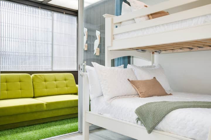 Svetlá a veselá spálňa s poschodovou posteľou s ostrou bielou posteľnou bielizňou, moderným stropným ventilátorom a štýlovými posuvnými dverami skrine. Izba je zdobená živými tropickými tapetami a jedinečnou kruhovou poličkou, ktorá vytvára zábavný a príjemný priestor.