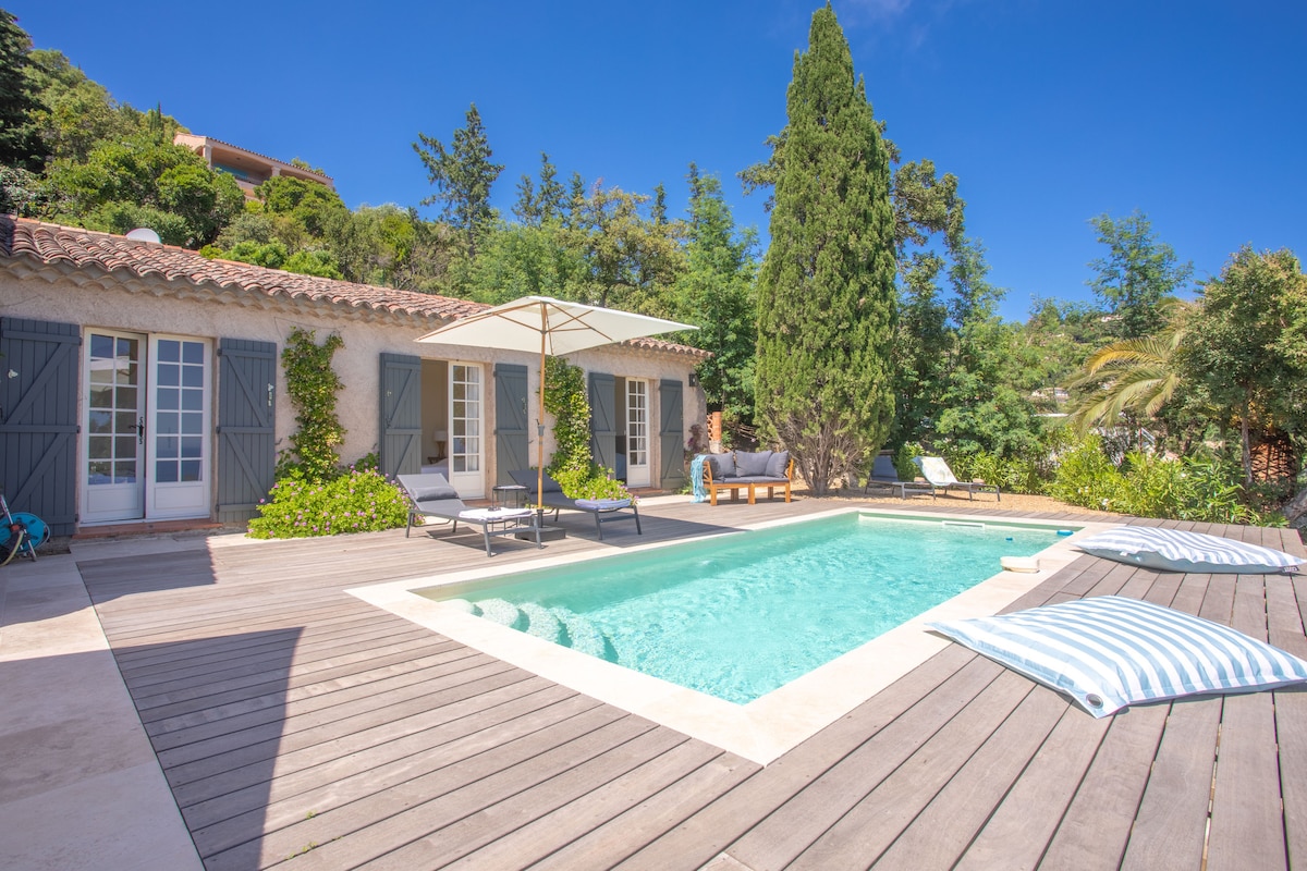 A private swimming pool is surrounded by a wooden deck, offering loungers and large cushions for relaxation. Lush greenery and tall trees frame the area, while a shaded seating area is visible in the background, providing a serene outdoor space with unobstructed views.