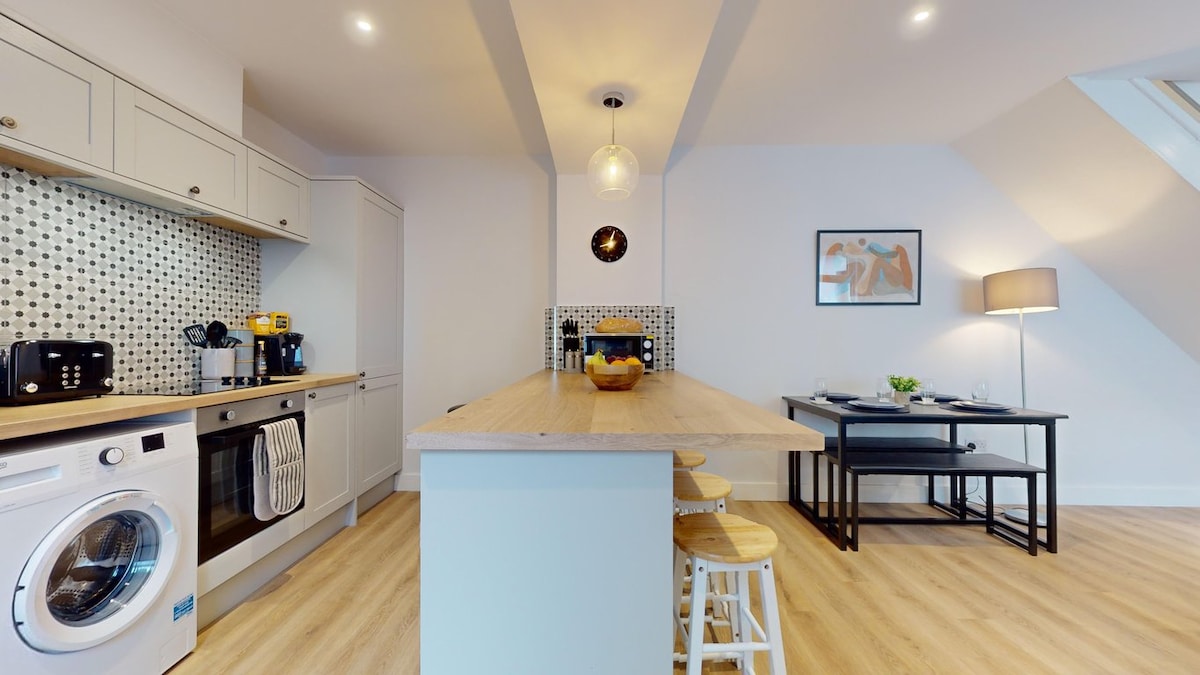 A modern kitchen area features a long wooden island with seating, surrounded by sleek cabinetry and appliances. A round pendant light hangs above, while a dining table with chairs is positioned nearby, illuminated by soft, natural light from overhead windows.