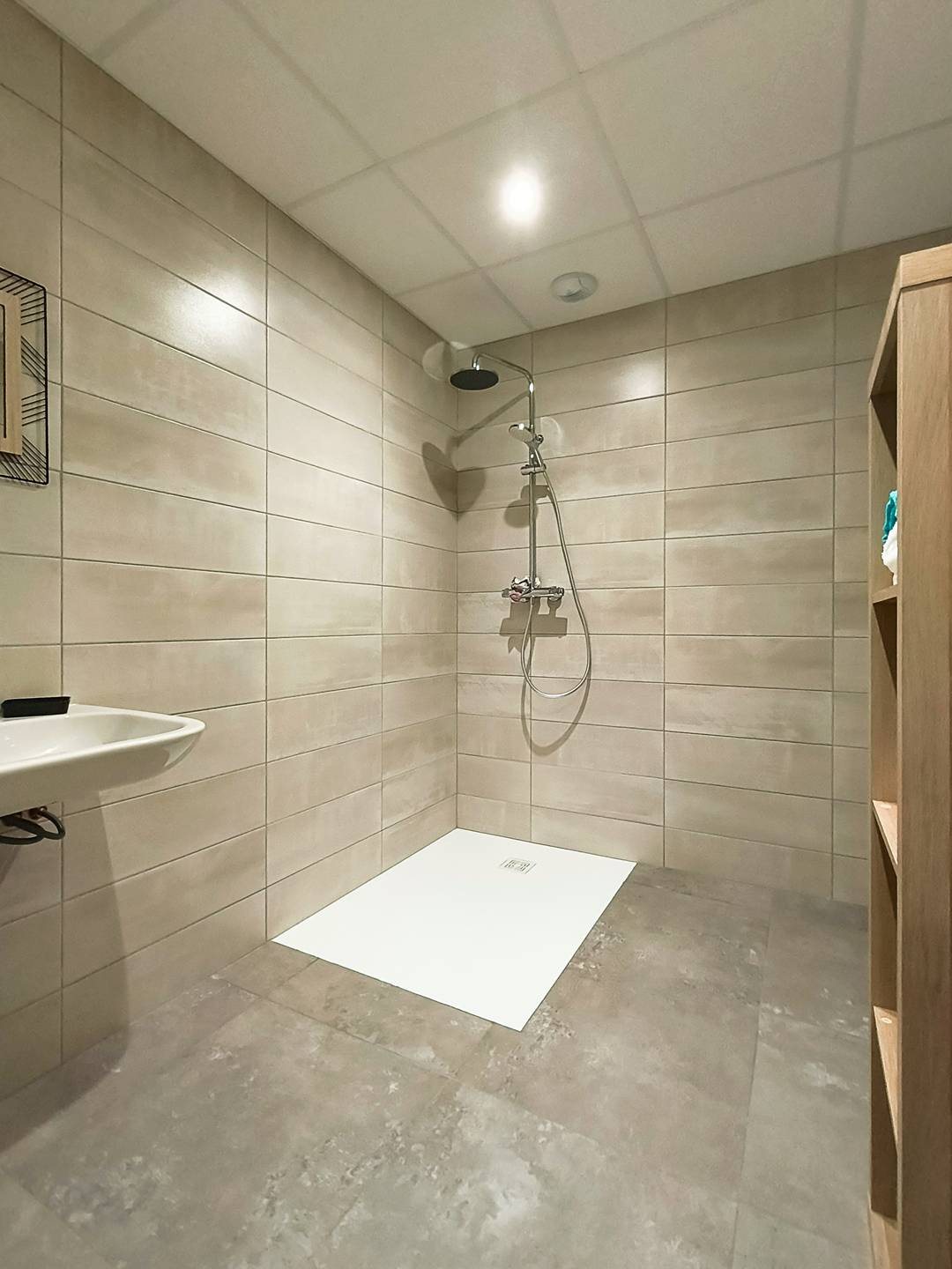 A well-designed bathroom features a spacious shower area with textured tiles lining the walls. A modern shower fixture is mounted above a sleek, white shower tray, while a minimalistic sink is positioned to the left. Shelving holds neatly arranged towels, contributing to an organized aesthetic.