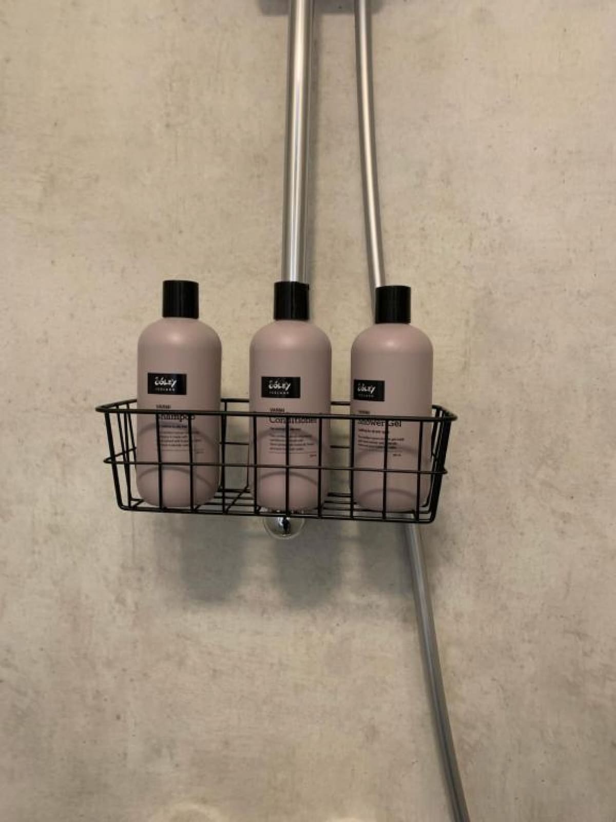 A metal shower caddy is attached to the wall, holding three bottles of personal care products. The bottles are uniformly shaped with black caps and feature minimalistic labels. The wall behind is finished in a light, textured concrete, enhancing the modern aesthetic.