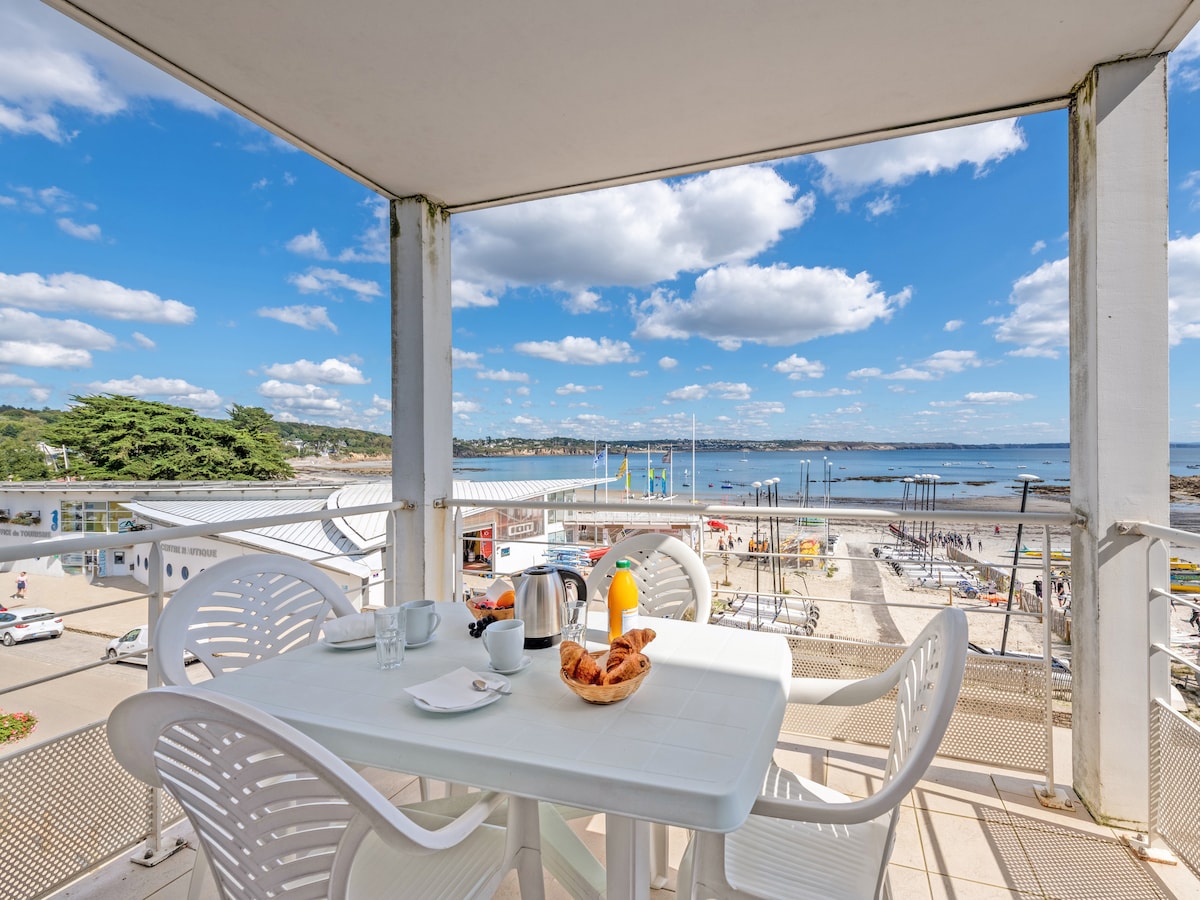 A balcony dining area is shown, furnished with a white table and four matching chairs. A small bowl of croissants sits alongside a coffee pot and juice, all set against a scenic view of the beach and sea under a blue sky with scattered clouds.