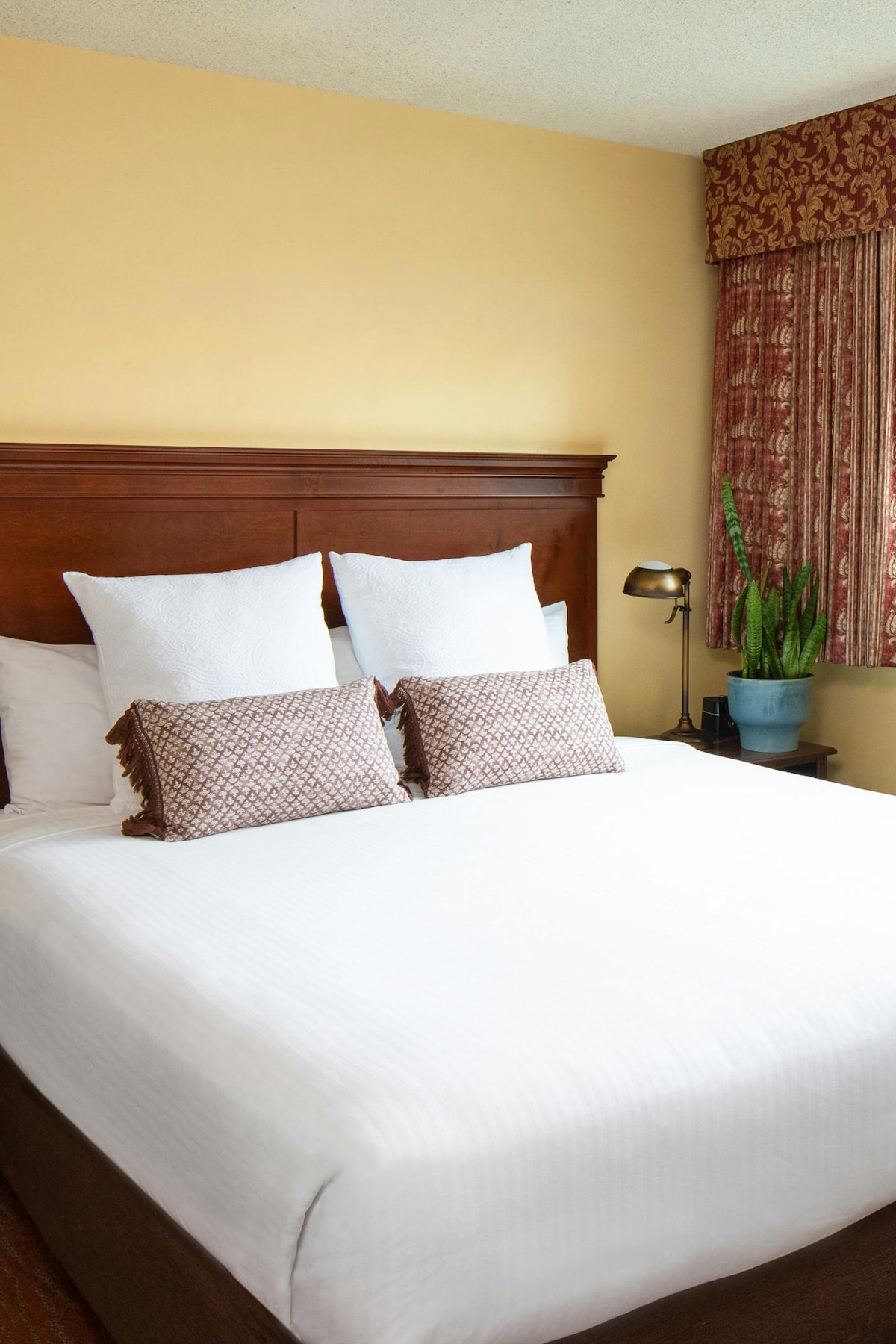 A cozy sleeping area is presented with a spacious bed featuring crisp white linens and decorative throw pillows. A wooden headboard adds warmth, while a bedside lamp illuminates the space. A potted plant is positioned nearby, complementing the soft hues of the room.