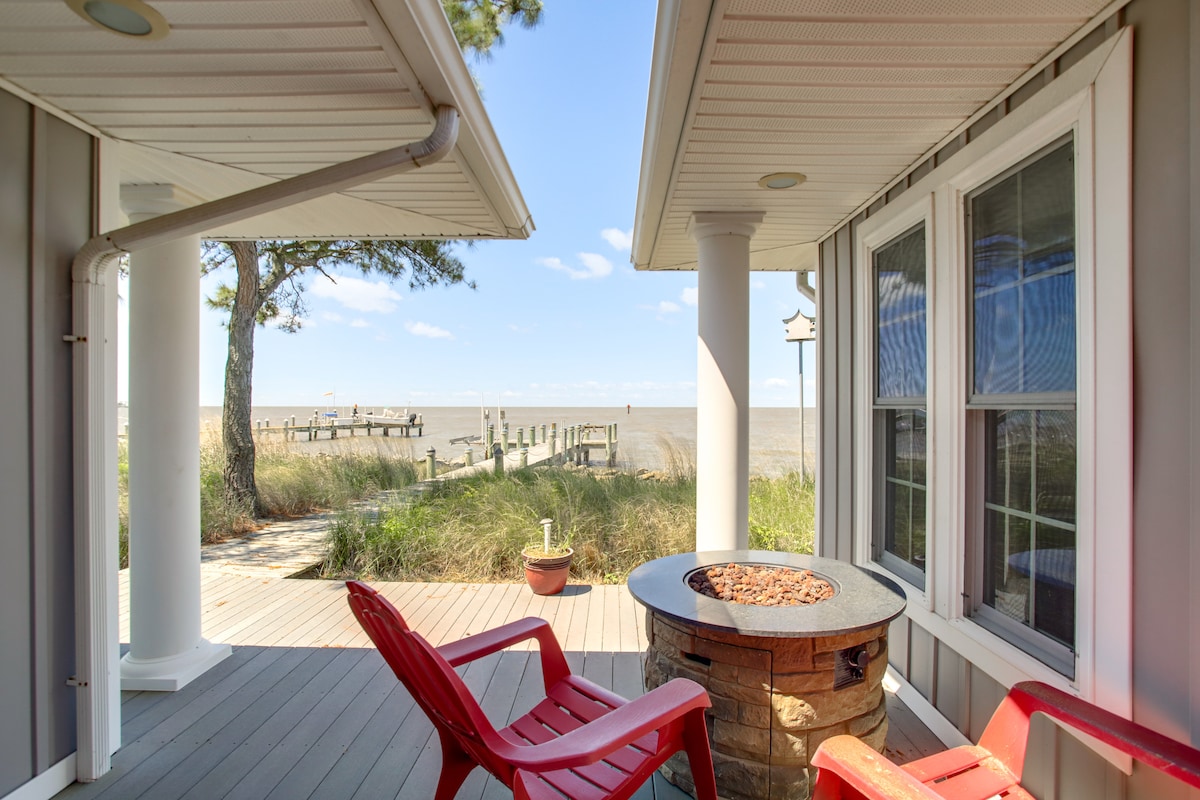 A porch area is showcased, featuring two red chairs positioned around a stone fire pit. The view extends to a private dock across a bay, surrounded by grassy landscape, under a clear sky.
