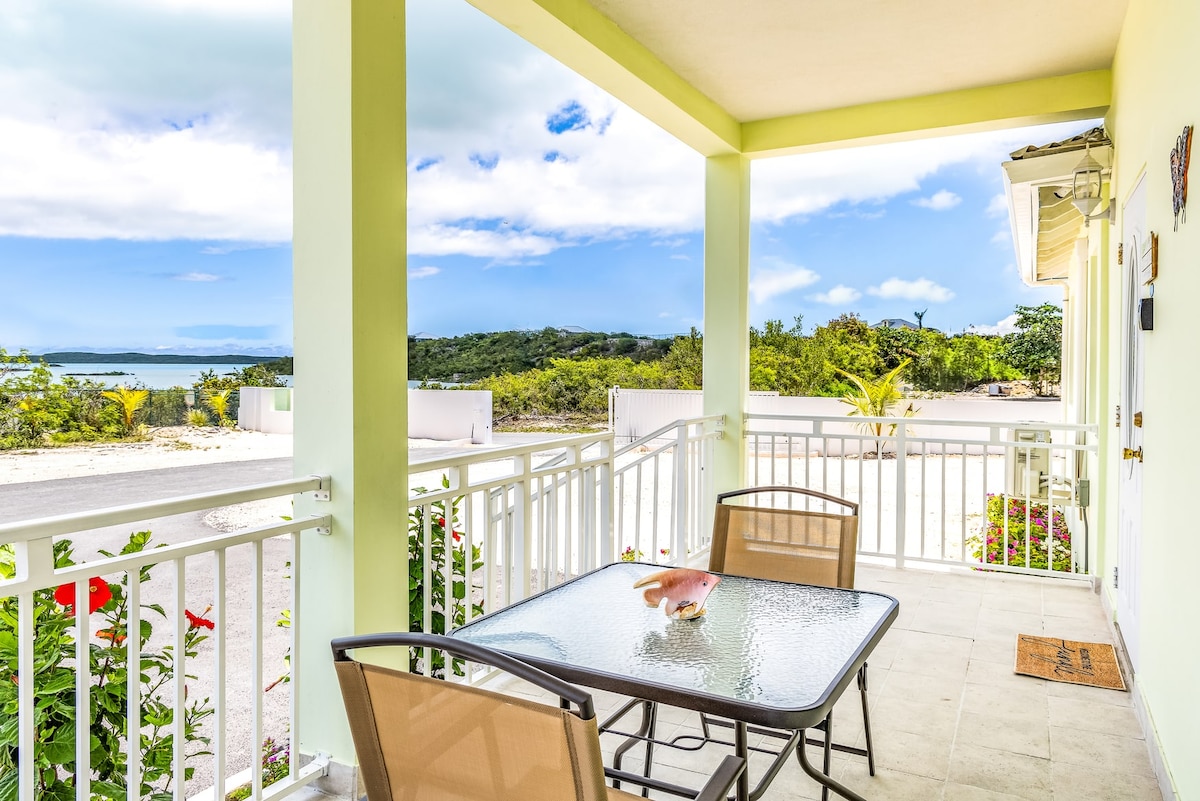 A private front porch features a glass-top table surrounded by four metal chairs. The space offers views of lush greenery and the tranquil Chalk Sound lagoon in the distance, framed by a bright blue sky with scattered clouds.