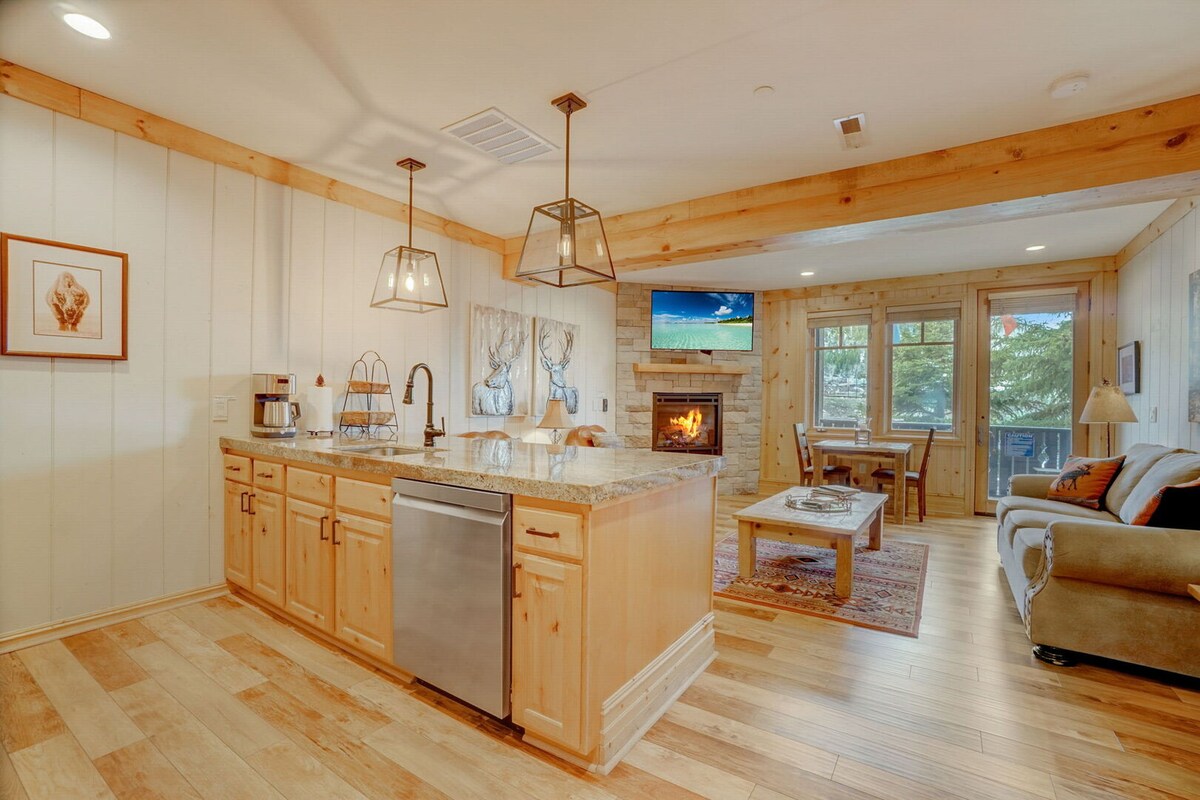 An inviting open-concept living area features a well-equipped kitchen with wooden cabinetry and modern appliances. A cozy seating area with a sofa is positioned near a stone fireplace. Warm wood accents and natural light enhance the atmosphere, while a dining space with a table and chairs is visible.