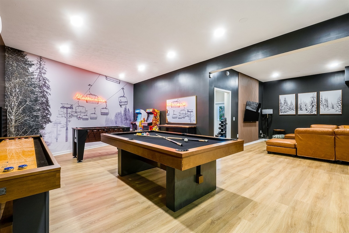 A recreation room is displayed, featuring a pool table, foosball table, and shuffleboard, accompanied by retro arcade games. A stylish brown leather sectional sofa provides seating, while wall art depicting snowy trees adds a cozy touch. Soft lighting enhances the inviting atmosphere.