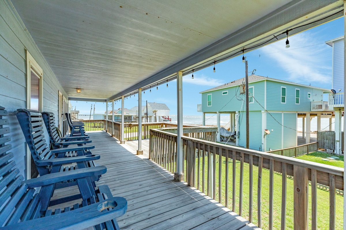 A spacious wraparound deck is featured, lined with several blue chairs facing the ocean. Sunlight brightens the area, and views of the nearby homes and greenery are visible. String lights hang above, enhancing the inviting outdoor space.