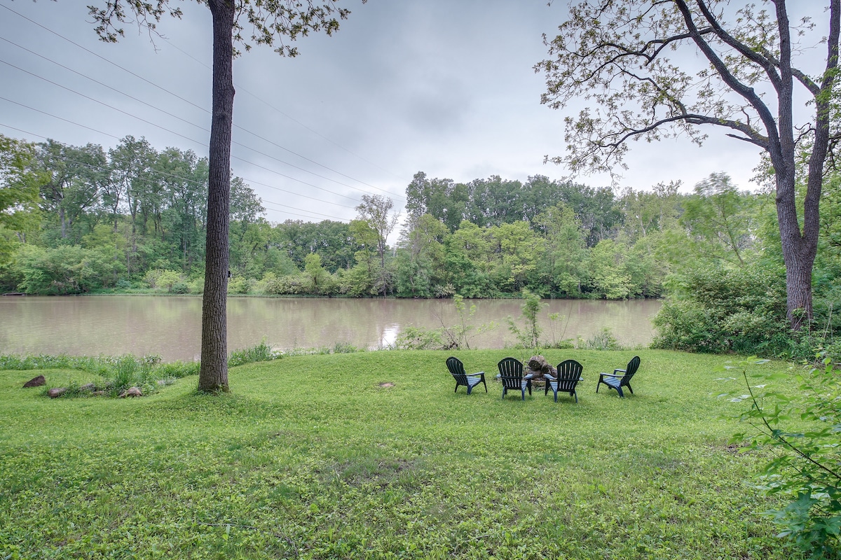 A serene outdoor space featuring a grassy area adjacent to the Scioto River. Four black Adirondack chairs are positioned in a circle under the shade of trees, providing a peaceful spot for relaxation and conversation with nature.