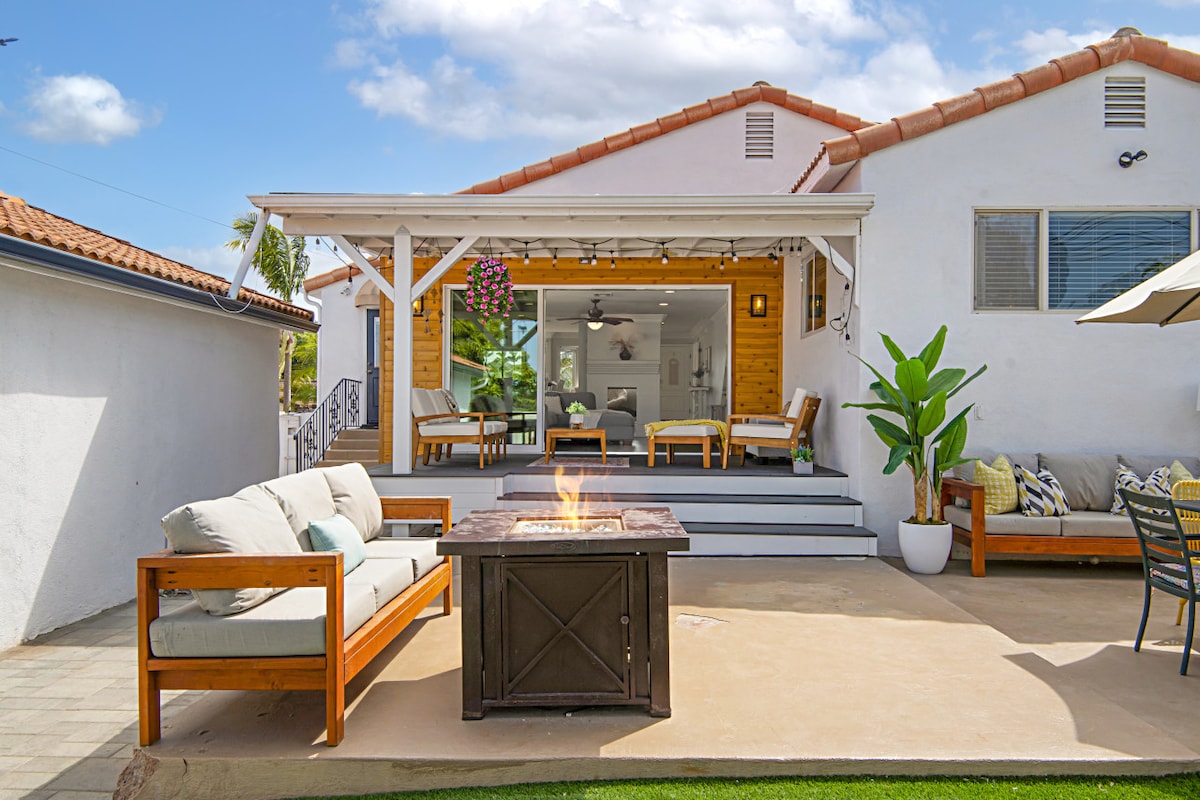 An inviting outdoor living space showcases a covered patio with comfortable seating and a central fire pit. Adjacent to the patio is a well-maintained lawn, surrounded by potted plants and a tranquil atmosphere, perfect for relaxation and gatherings.