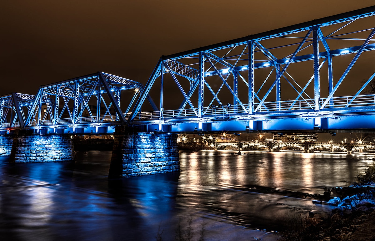 An illuminated blue bridge spans across a calm waterway under a night sky. The reflections of the lights create a serene atmosphere, with gentle ripples visible on the water's surface. The sturdy brick pillars support the bridge, enhancing its architectural beauty.