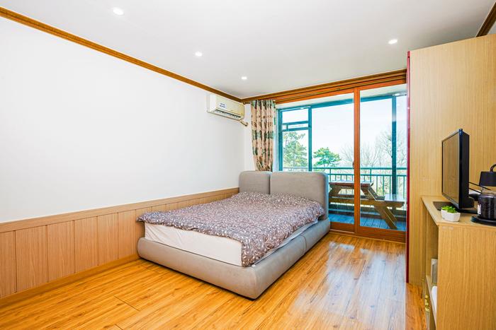 A spacious room is furnished with a large bed featuring a neutral-colored coverlet. Natural light fills the space through a large window, which offers a view of the surrounding area. A small television and a storage unit can be seen along the wall.