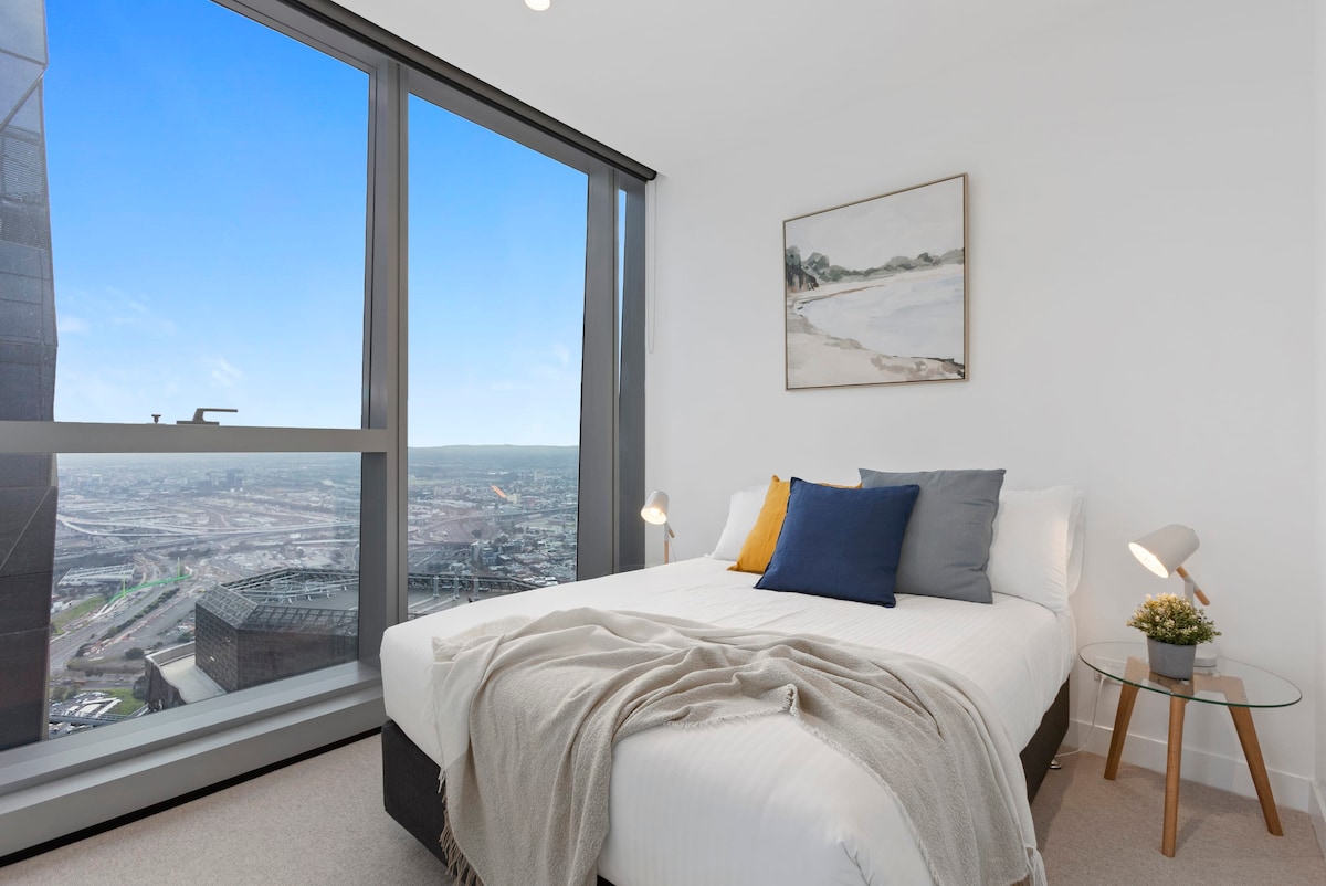 A cozy bedroom features a spacious bed adorned with neutral bedding and accent pillows in blue and yellow. Large windows allow natural light to fill the room, offering sweeping city views. A small side table with a lamp complements the elegant interior design.