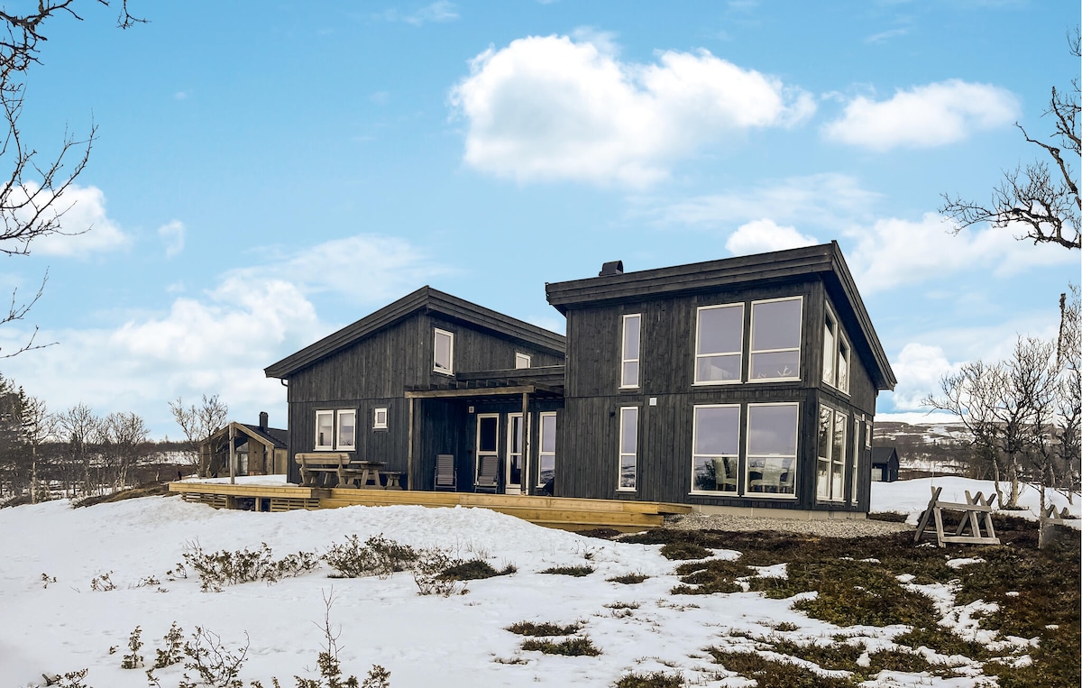 The holiday home is situated on a snow-covered landscape, featuring a dark wooden exterior and large panoramic windows. A spacious terrace is visible, accompanied by outdoor seating options. The surrounding area showcases sparse vegetation and a clear blue sky, contributing to a serene atmosphere.