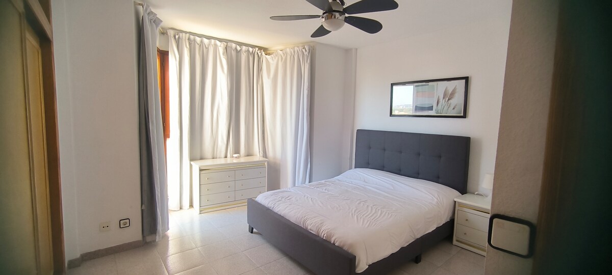 A spacious bedroom features a large bed with a tufted gray headboard, complemented by a white bedspread. Two curtains frame a window, allowing natural light to fill the room. A chest of drawers stands against the wall, while a ceiling fan provides additional airflow.
