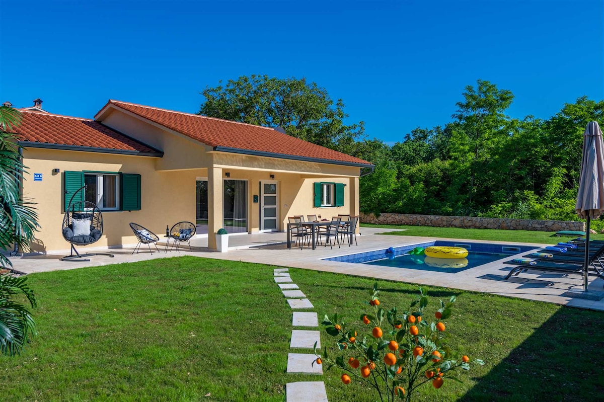 A single-story villa is presented with a bright yellow exterior and a terracotta roof. Outdoor seating is arranged near the pool, along with sunbeds and an umbrella. A patch of greenery features vibrant orange trees, while the surrounding lush foliage creates a serene atmosphere.