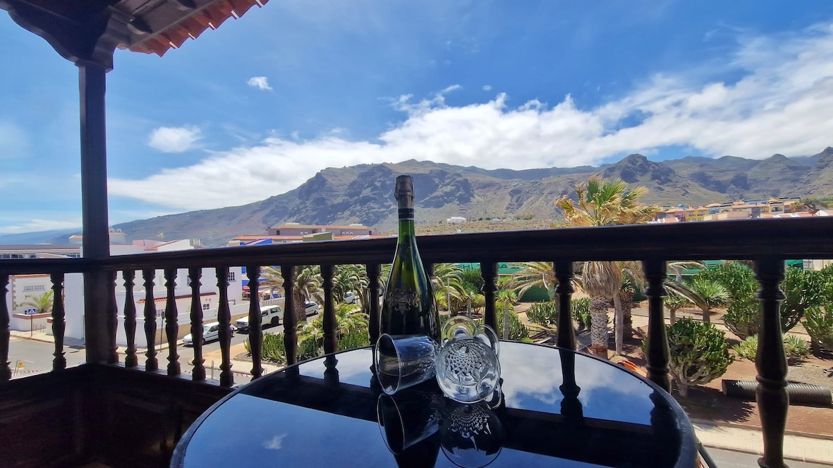 A balcony table is set with a bottle of sparkling wine and a glass. Lush greenery surrounds the area, with mountainous views in the background under a bright blue sky. The wooden railing adds a rustic touch to the serene setting.