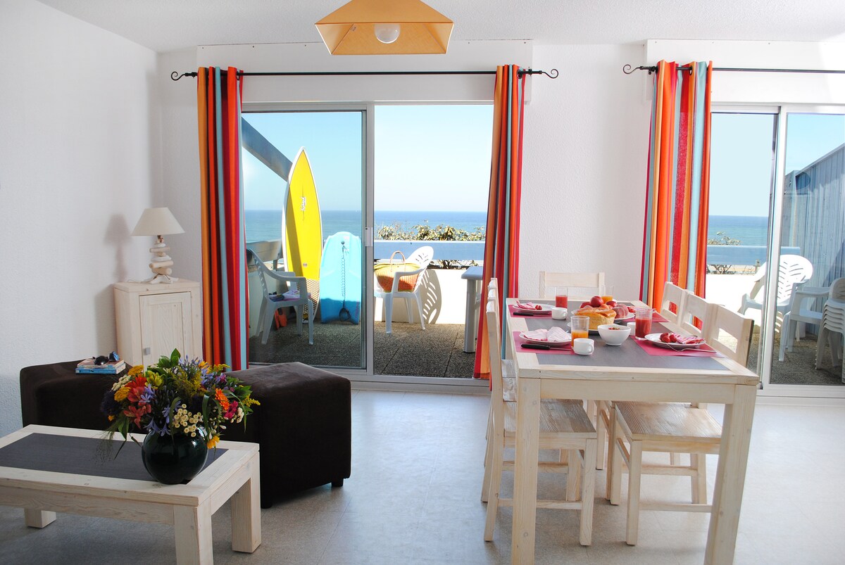 A bright living area features large glass doors that offer a view of the ocean. A dining table is set for four, accompanied by a cozy sofa adorned with flowers. Vibrant curtains frame the space, while beach gear is visible on the terrace outside.