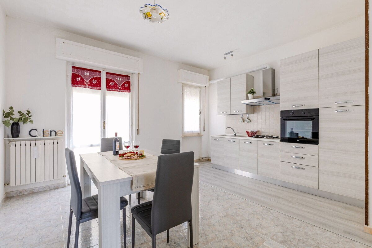 A modern kitchen is equipped with built-in appliances and sleek cabinetry. A dining table with four chairs is positioned near a window, allowing natural light to fill the space. Decor elements include potted plants and kitchen accessories, creating a welcoming atmosphere.
