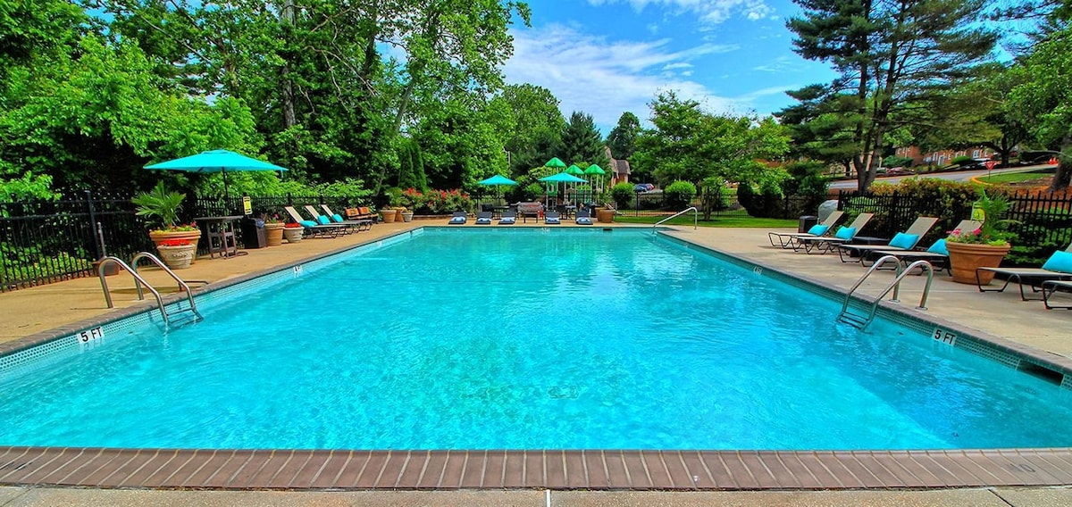 The outdoor pool area is surrounded by green trees and vibrant landscaping. Lounge chairs are positioned around the pool, along with tables and umbrellas for shade. A clear blue sky is visible above, adding to the inviting atmosphere of the space.