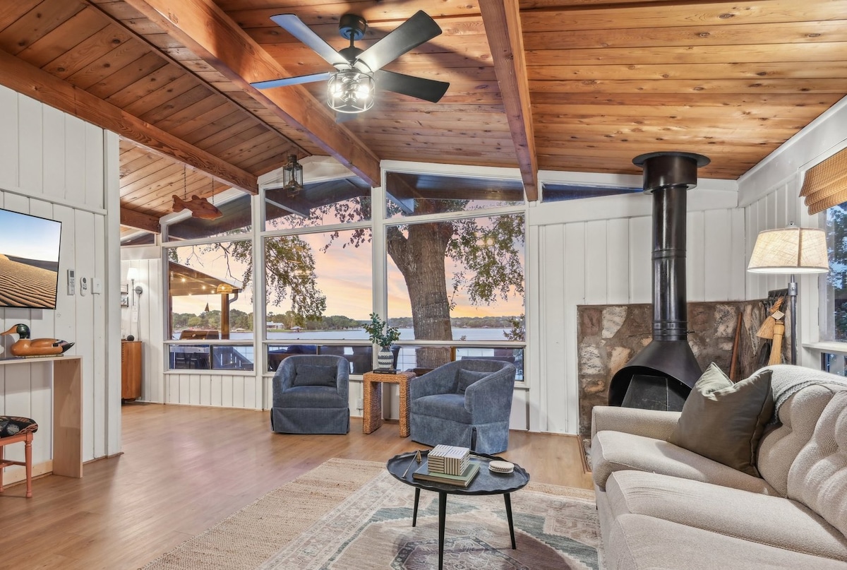 The open living area features large windows that frame views of the lake at sunset. A comfortable sofa and two swivel chairs are arranged around a low coffee table, while warm wood beams add character to the ceiling. A modern ceiling fan and decorative fireplace enhance the ambiance.