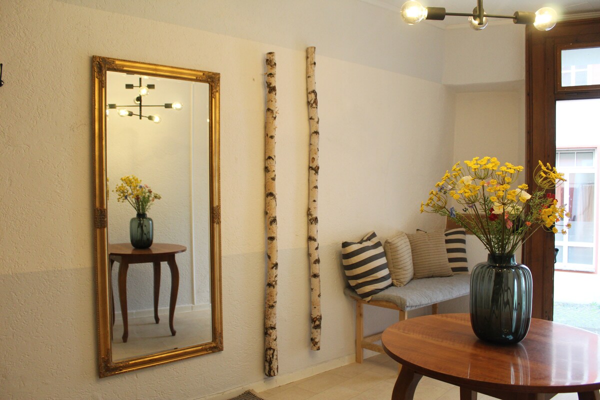 A welcoming entrance showcases a large mirror with a decorative gold frame, reflecting light within the space. A round wooden table is positioned beside a seating area adorned with striped cushions. A vase filled with fresh flowers adds a touch of color to the neutral decor.