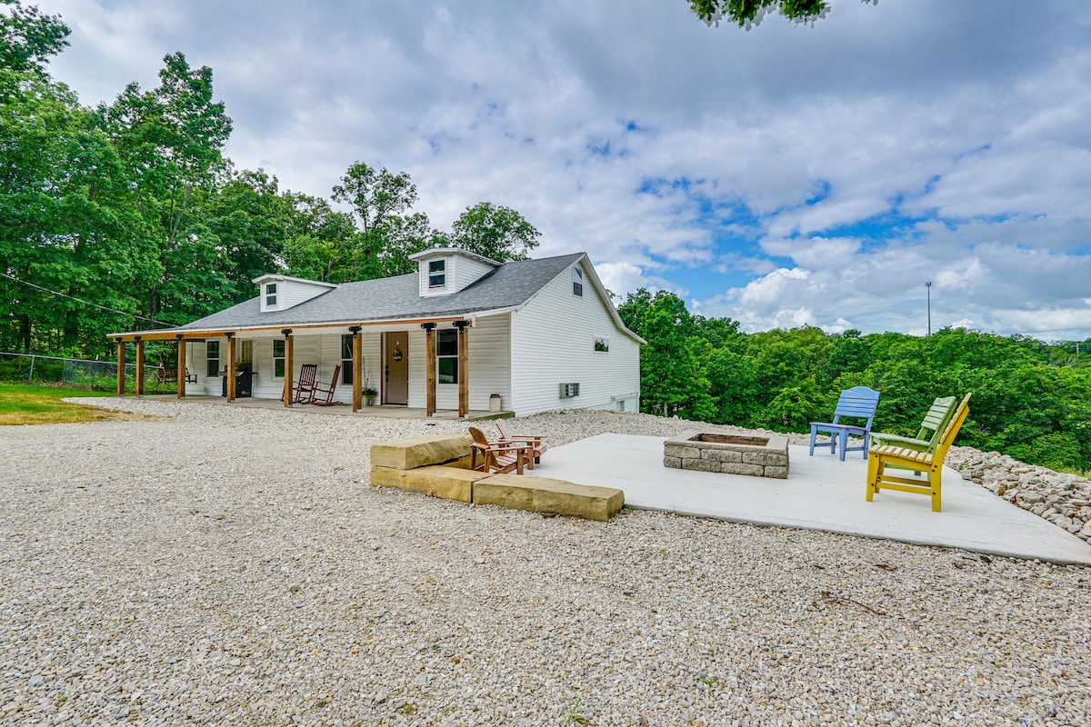 A view of the cabin shows a spacious gravel area in front, with a mix of seating options including rocking chairs and colorful chairs surrounding a stone fire pit. Lush trees provide a natural backdrop, enhancing the outdoor space.