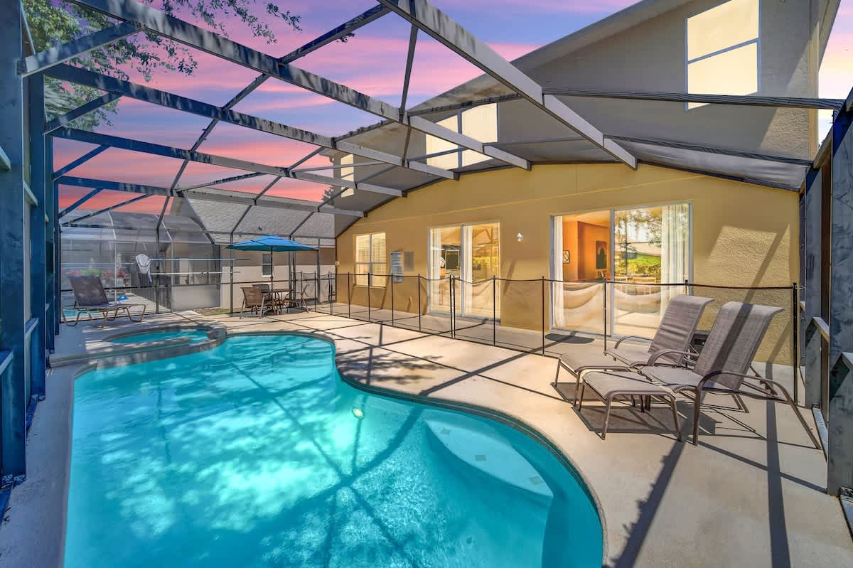 A private pool area is enclosed by a screened lanai, featuring a curved pool with lounge chairs positioned along its edge. Large sliding glass doors provide access to the home, while a serene exterior is enhanced by a colorful sky at dusk.