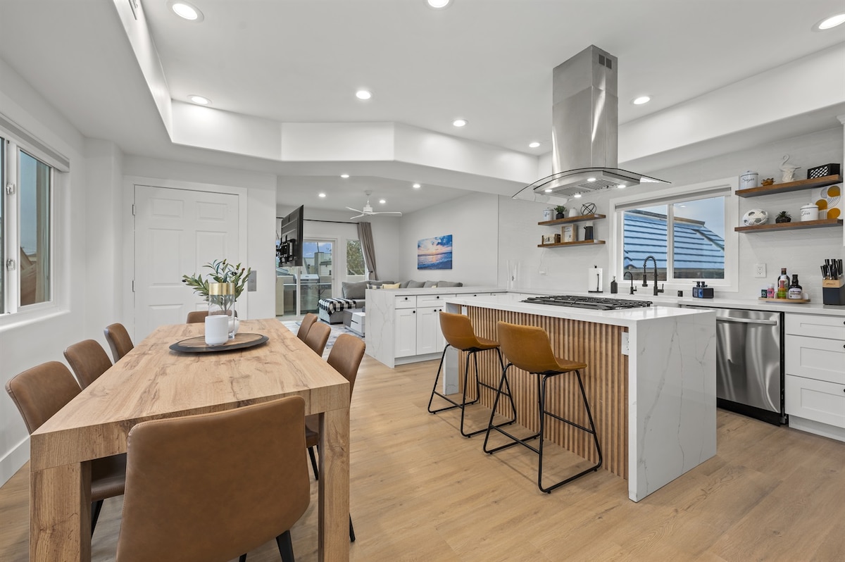 A modern kitchen and dining area features a large wooden dining table surrounded by brown upholstered chairs. A kitchen island with bar seating and a stainless-steel range hood is visible, complemented by sleek cabinetry and ample natural light from windows.