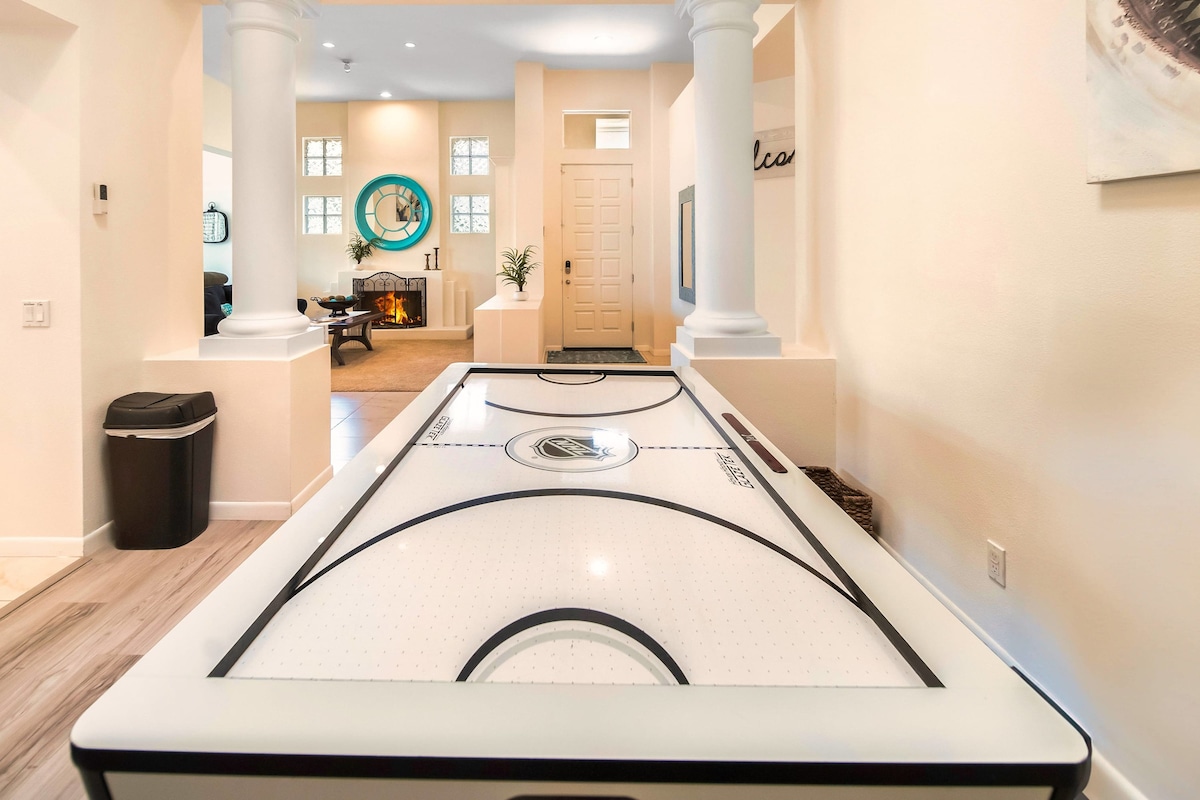An air hockey table is positioned in a well-lit hallway, serving as a point of entertainment. Columns frame the entrance to a spacious living area, where natural light pours in through multiple windows, creating an open and inviting atmosphere.