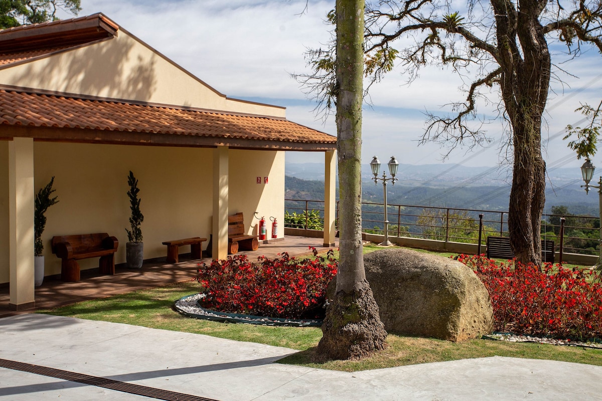An exterior view of a building featuring a terracotta roof and light-colored walls. Surrounding the entrance are neatly arranged plants and vibrant red flowers. A large boulder and wooden benches are positioned nearby, with a scenic landscape visible in the background.