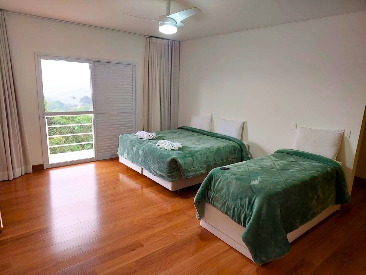 A tranquil bedroom features two spacious beds, each covered with soft green blankets. Natural light filters in through the sliding door, which leads to a balcony with views of the surrounding landscape. Light wood flooring complements the calming atmosphere.