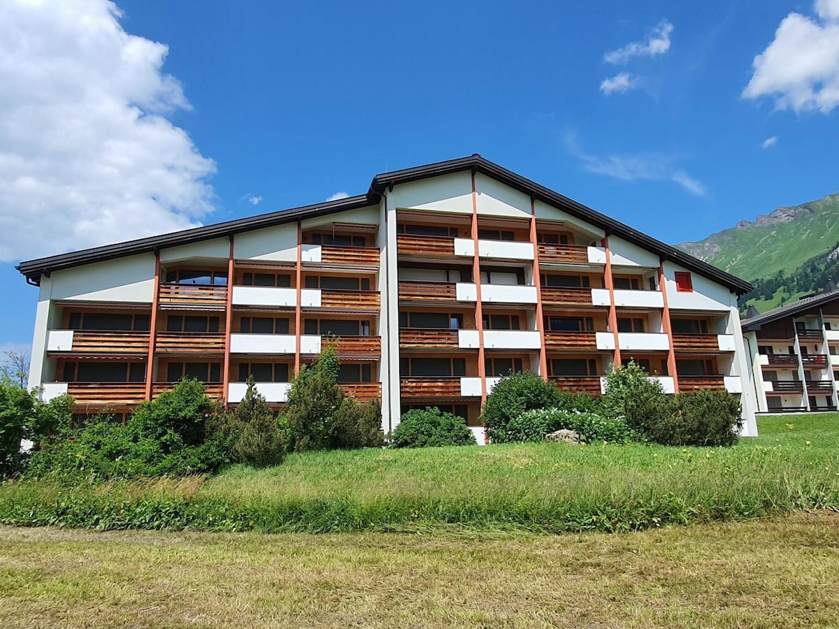 A three-story apartment building is set against a backdrop of greenery and mountains. Balconies with wooden railings are visible on each level, framed by surrounding grass and shrubs under a blue sky with scattered clouds.