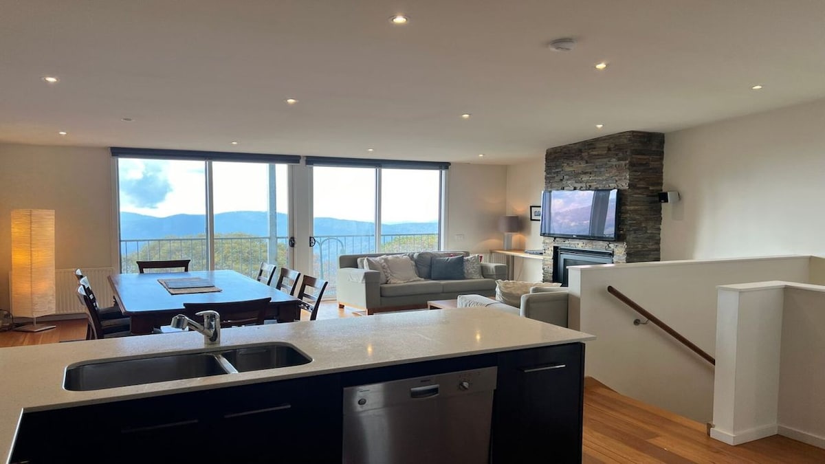 A modern open-concept living area features large windows offering expansive mountain views. Polished wooden floors enhance the space, which includes a designated dining area with seating for eight and a cozy seating arrangement by a stone fireplace.