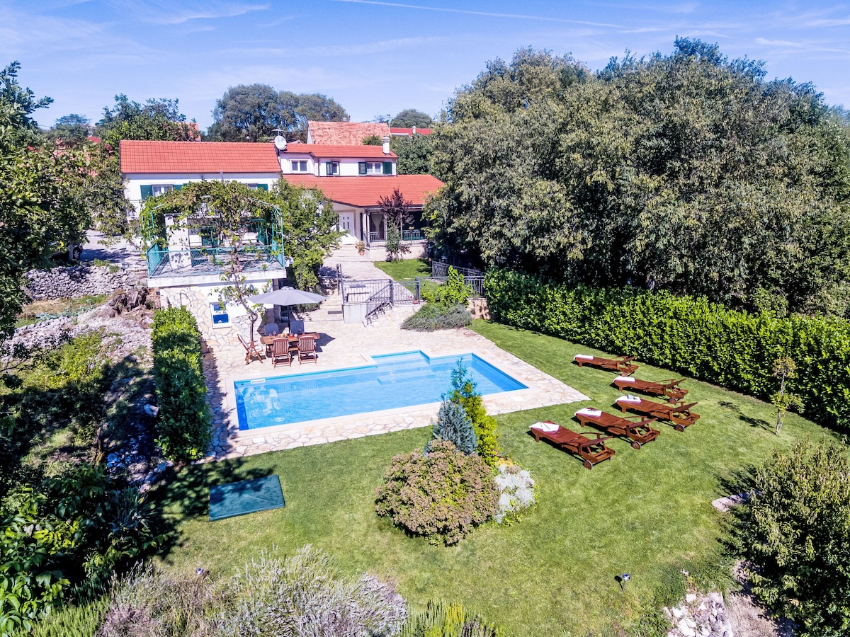 An aerial view captures the spacious outdoor area, featuring a rectangular swimming pool surrounded by a stone patio. Comfortable lounge chairs are positioned nearby on the lush lawn. The house, with red roof tiles, is in the background, bordered by trees and greenery, enhancing privacy.