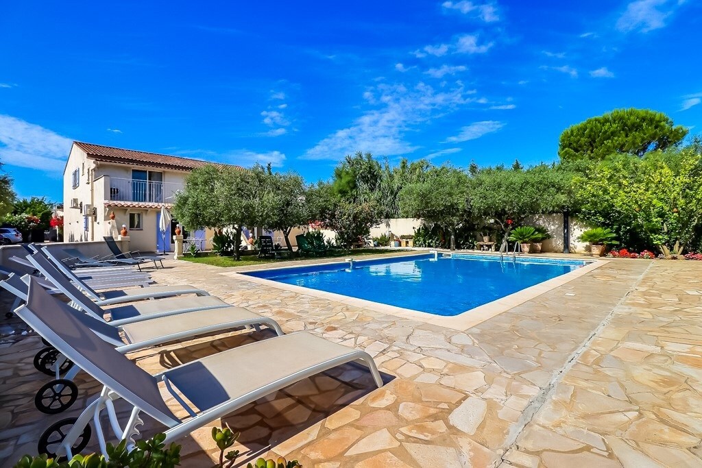 A spacious outdoor area is featured, showcasing a large swimming pool surrounded by sun loungers. Lush greenery and trees provide shade, creating a pleasant atmosphere. The flat is visible in the background, while a clear blue sky enhances the inviting setting.