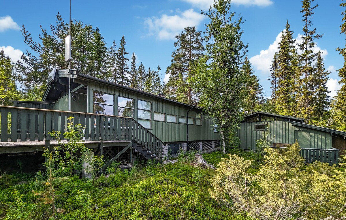 The exterior of a green wooden house is shown, nestled among lush greenery and towering trees. A deck extends from the building, providing a pathway to the entrance. The overall design features large windows, allowing natural light to illuminate the interior spaces.