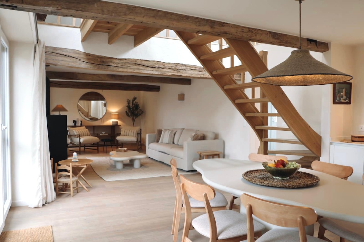A spacious living area is characterized by a blend of modern and rustic elements. Wooden beams accent the ceiling, while a staircase leads to the upper level. A light-colored sofa and coffee table are arranged, accompanied by a dining table surrounded by wooden chairs.