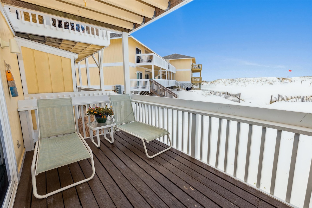 A private deck is shown featuring two light-colored lounge chairs positioned near a small table with a potted flower arrangement. The view beyond includes sandy dunes and a clear blue sky, hinting at nearby beach access.