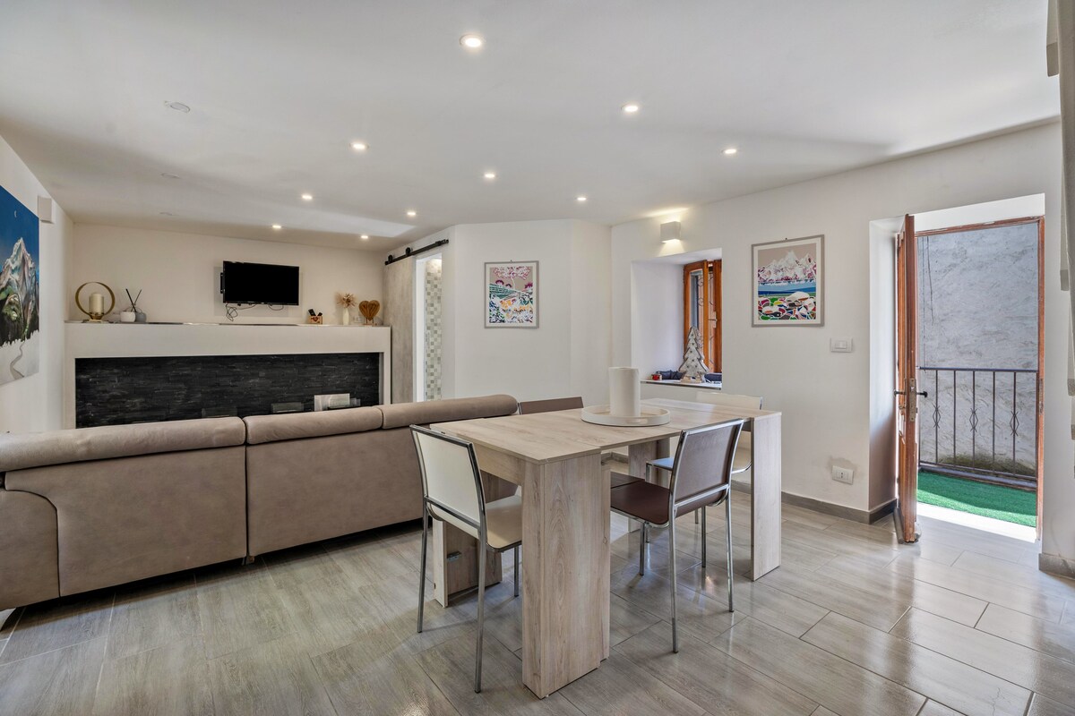 A modern living area features a spacious sofa facing a wall-mounted television. A dining table with several chairs is situated nearby, with natural light streaming through doors that lead to a private balcony. The tile flooring is complemented by neutral-colored walls.