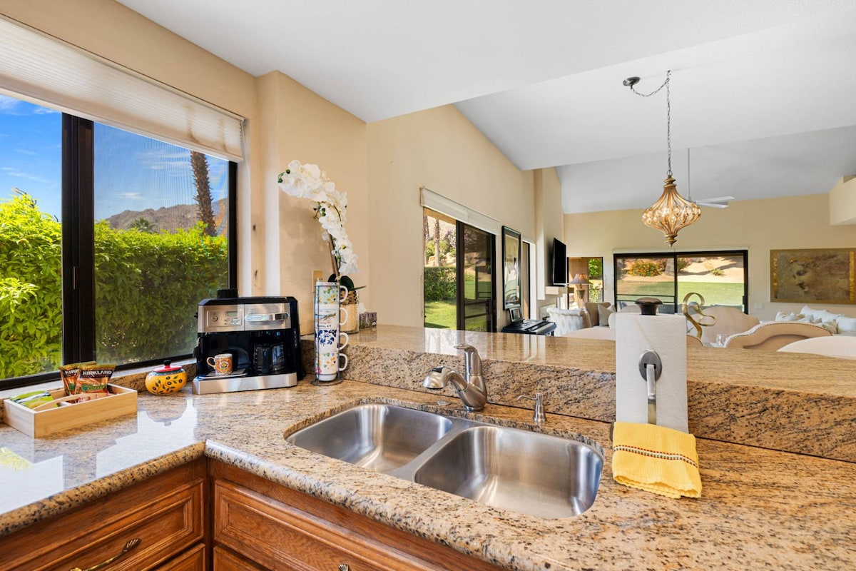 A newly remodeled kitchen is showcased with stainless steel appliances and granite countertops. A large window allows natural light to fill the space and offers views of the outdoor greenery. Decorative elements include a coffee maker and an arrangement of flowers.