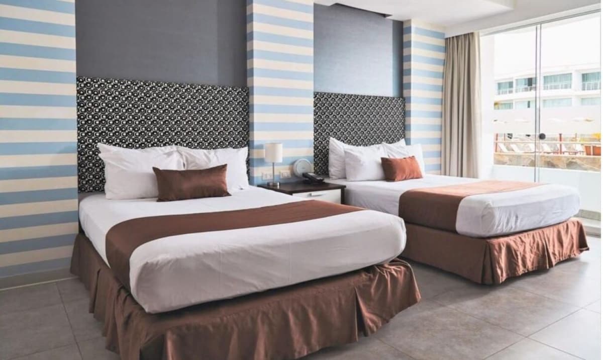 A bright and spacious bedroom features two comfortable beds with coordinating brown bed skirts. Decorative headboards enhance the room's design, while soft natural light fills the space through large windows with sheer curtains. A bedside table and lamp provide functionality.