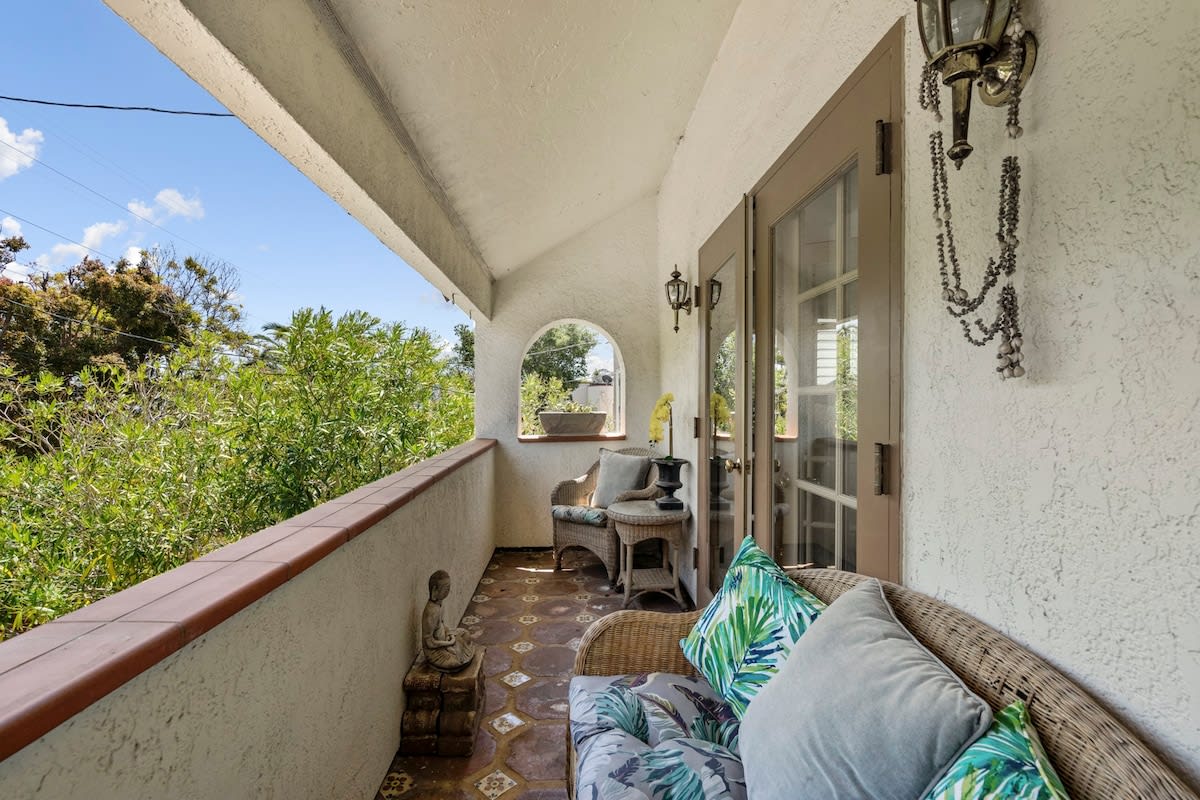 An outdoor patio area features comfortable seating with decorative cushions. The space is framed by arches, allowing views of surrounding greenery. Two light fixtures add subtle illumination, while a small statue adds a decorative touch alongside the seating. Natural light highlights the tiled flooring.