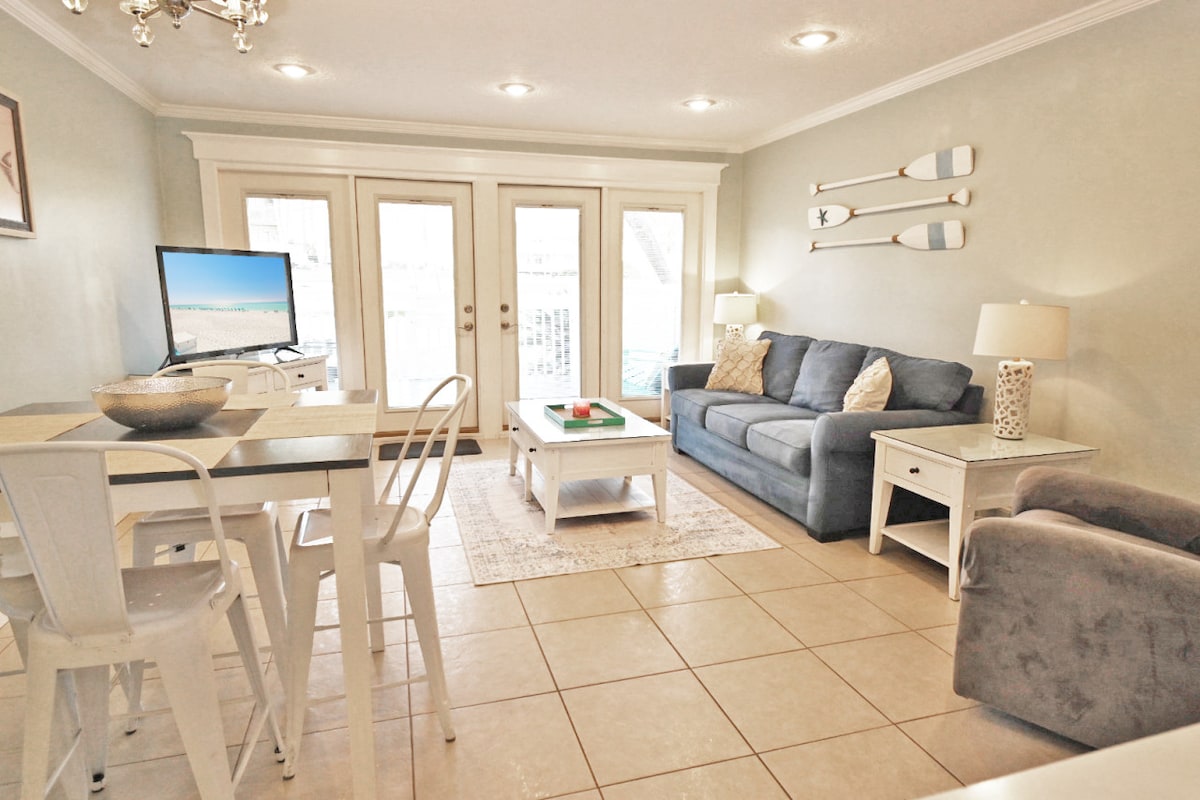 The living and dining area features a soft blue sofa and a rocking recliner. A flat-screen TV is positioned across from a dining table with four white chairs. Natural light filters in through double glass doors leading to an outdoor space.